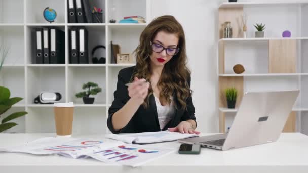 Attractive purposeful stylish modern businesswoman working at the laptop simultaneously writing something into report and browsing information on her mobile in office - Materiaali, video