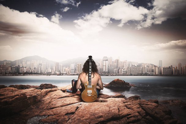 Vintage gitarzysta siedzi na skałach brzegu patrząc na miasto w dzień labiryntu  - Zdjęcie, obraz