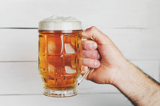 Male hand holding a full beer mug with foam on white wooden background. Toned image. - Photo, image