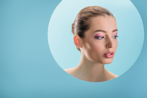 portrait of beautiful naked woman with glossy lips, pink eyeshadow looking away through paper round hole isolated on blue - Photo, Image