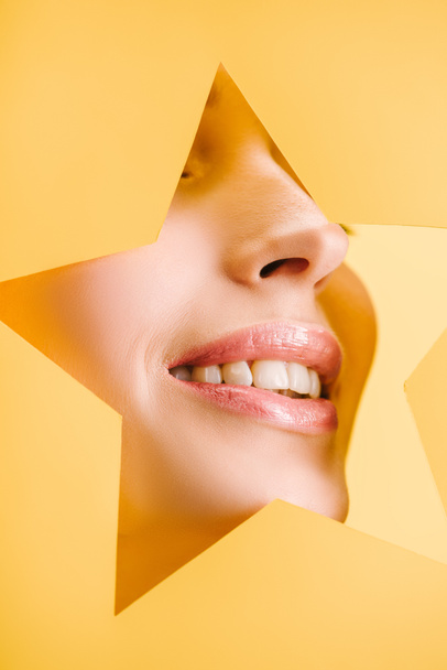 abgeschnittene Ansicht der schönen Frau mit glänzenden Lippen in Papier sternförmigen Loch lächelt isoliert auf gelb - Foto, Bild