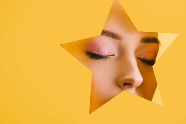 retrato de mujer hermosa con maquillaje brillante en el agujero en forma de estrella de papel con los ojos cerrados aislados en amarillo
 - Foto, imagen