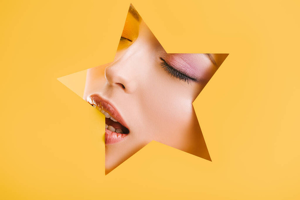 portrait of beautiful woman with shiny makeup in paper star shaped hole with closed eyes and open mouth isolated on yellow - Фото, изображение