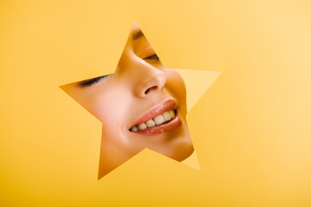 portrait of beautiful woman with shiny makeup in paper star shaped hole smiling isolated on yellow - Photo, Image