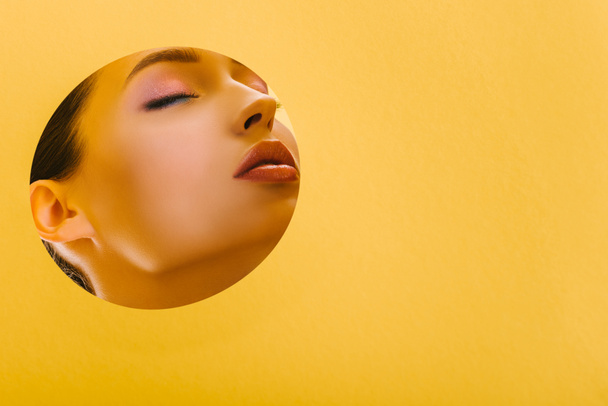 portrait of beautiful woman with shiny makeup in paper round hole with closed eyes isolated on yellow - Photo, Image