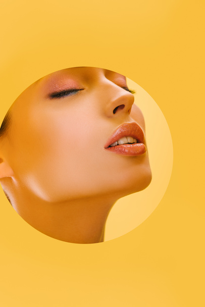 portrait of beautiful woman with shiny makeup in paper round hole with closed eyes isolated on yellow - Zdjęcie, obraz