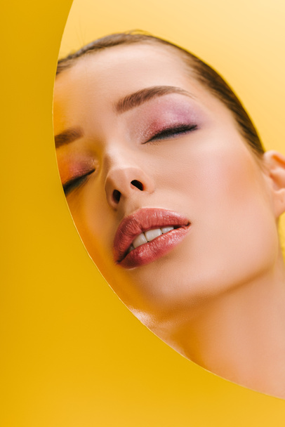 retrato de mujer hermosa con maquillaje brillante en papel agujero redondo con los ojos cerrados aislados en amarillo
 - Foto, imagen