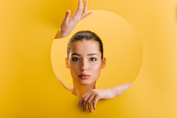 portrait of beautiful woman with shiny makeup in paper round hole with hands isolated on yellow - Valokuva, kuva