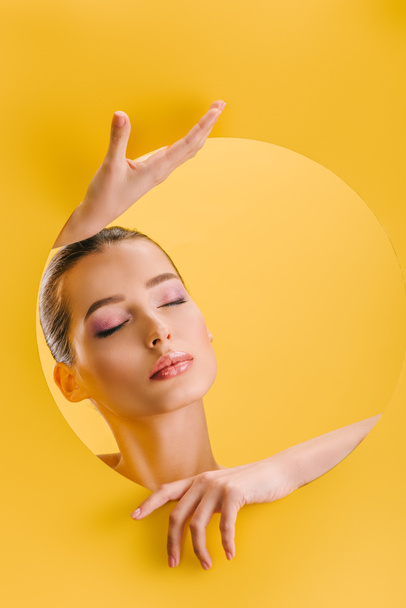portrait of beautiful woman with shiny makeup in paper round hole with hands and closed eyes isolated on yellow - Foto, immagini