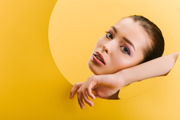 portrét krásné ženy s lesklým make-upem v papírové kruhové dírce s rukou izolovanou na žluté - Fotografie, Obrázek