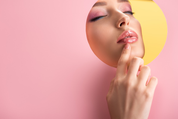 portrait of beautiful woman with shiny makeup in pink paper round hole with hand and closed eyes isolated on yellow - Фото, изображение