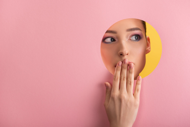 portrait of beautiful woman with shiny makeup in pink paper round hole covering mouth with hand isolated on yellow - Foto, imagen