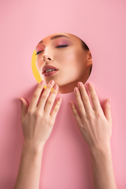 portrait of beautiful woman with shiny makeup and closed eyes in pink paper round hole with hands isolated on yellow - Foto, imagen