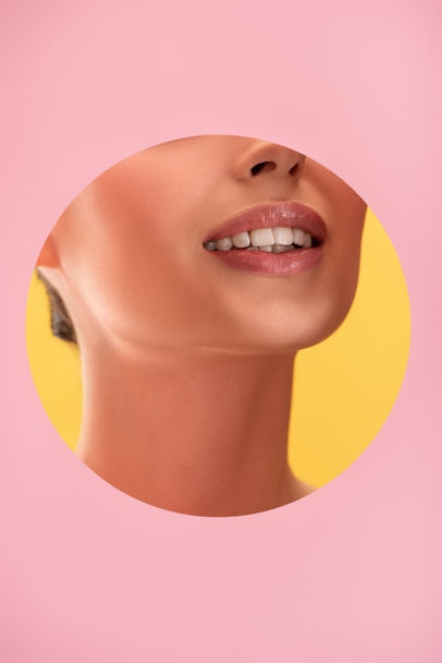 partial view of beautiful woman with shiny lips in pink paper round hole smiling isolated on yellow - Zdjęcie, obraz