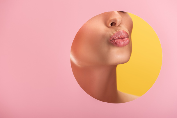 vue partielle de belle femme aux lèvres brillantes en papier rose trous ronds lèvres boudantes isolées sur jaune - Photo, image
