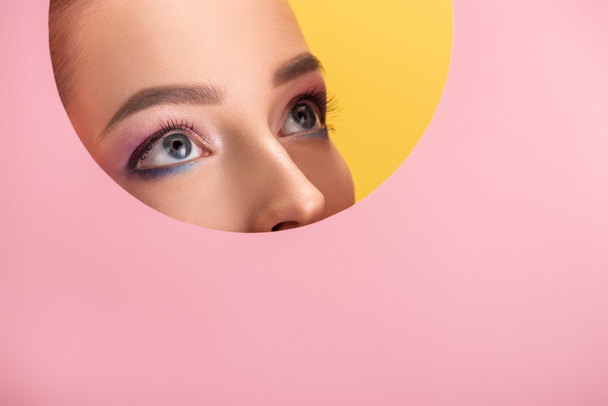 portrait of beautiful woman with shiny eyeshadow in pink paper round hole looking up isolated on yellow - Фото, зображення