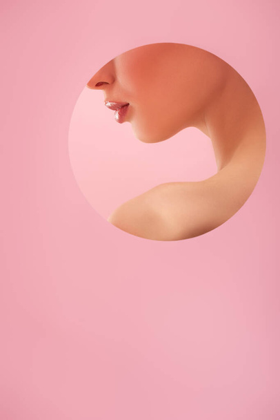 abgeschnittene Ansicht der schönen nackten Frau mit glänzenden Lippen in Papier runde Loch isoliert auf rosa - Foto, Bild