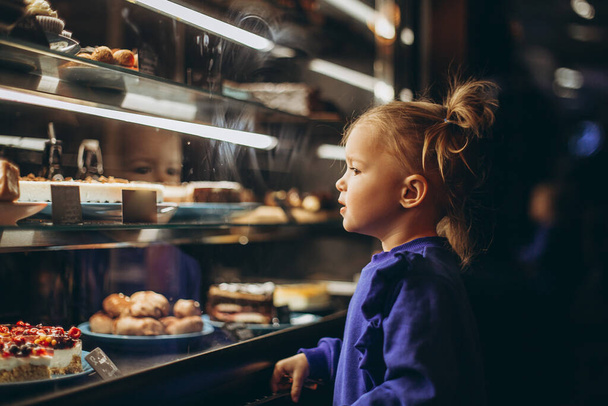 lovely girl admiringly examines a lighted showcase with various delicious cakes and desserts - Fotografie, Obrázek