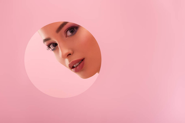 portrait of beautiful woman with shiny makeup in paper round hole isolated on pink - Фото, зображення