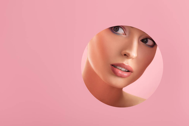 portrait of beautiful woman with shiny makeup in paper round hole isolated on pink - Foto, Imagen