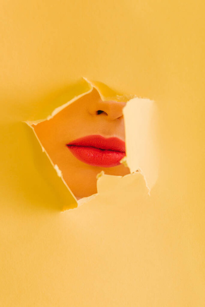 cropped view of beautiful woman with crimson lips in yellow paper torn hole - Fotografie, Obrázek