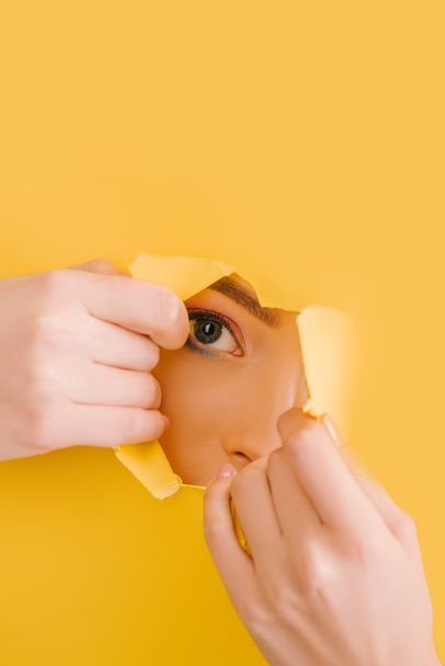 cropped view of beautiful woman looking through yellow paper torn hole - Фото, зображення