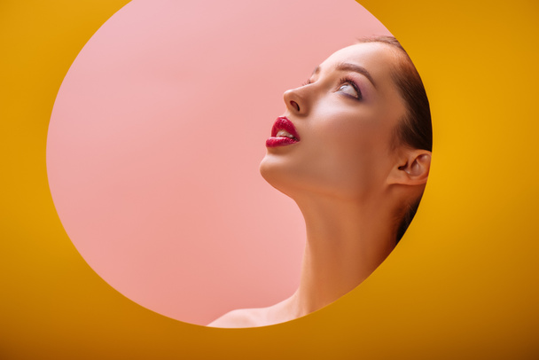 nackte schöne Frau mit purpurroten Lippen, die durch gelbes Papierkreisloch nach oben schaut - Foto, Bild