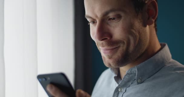 Happy guy using smartphone while standing near window - Felvétel, videó