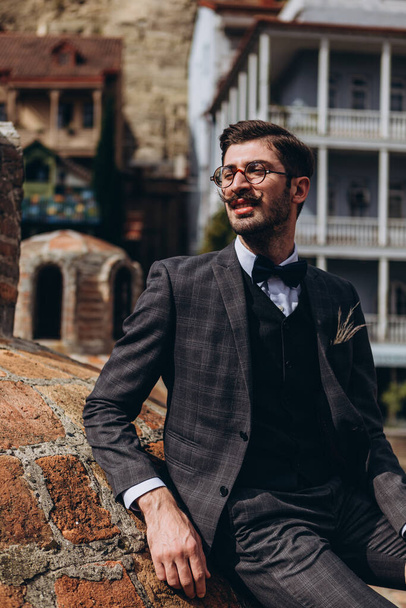 portrait of a stylish young Georgian man with mustache and vintage tuxedo posing for a photo on the background of the old city in Tbilisi - Fotó, kép