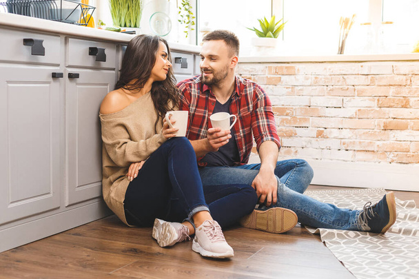 Lovely girl in casual sitting on the floor and talking with boyfriend. Young couple enjoying coffee or tea in kitchen. - Fotografie, Obrázek