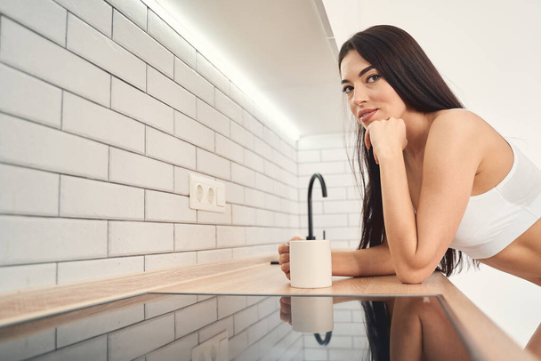 Smiling young woman bending over the kitchen bench holding cup of beverage - Фото, зображення