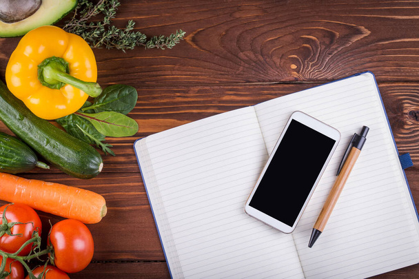 Čerstvá organická zelenina a ovoce. Pepř, rajče, avokádo, otevřený prázdný zápisník, telefon a pero na dřevěném pozadí. Zdravé jídlo a zdravý život koncept. Horní pohled - Fotografie, Obrázek