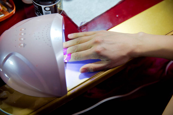 女性はマニキュアのトップカバーを硬化させるために鉛紫外線ランプに手を置きます。水平カラー写真。新鮮な美しいピンクの現代的なゲルポリッシュマニキュアと女性の手のクローズアップビュー. - 写真・画像