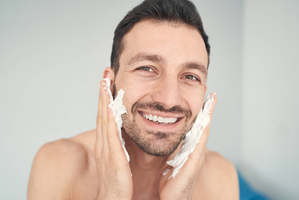 Joyful young man putting shaving soap on his cheeks - Foto, imagen