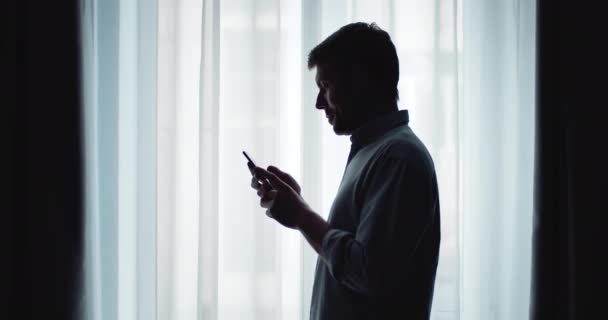 Side view of man standing near window and using smartphone - Felvétel, videó