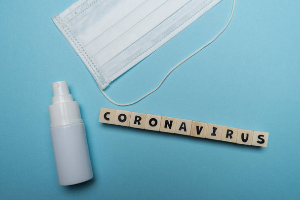 Higieniczna maska do twarzy z białą butelką do dezynfekcji rąk ze słowem Coronavirus napisanym na drewnianym bloku odizolowanym na niebieskim tle. Koncepcja zapobiegania wybuchom wirusa. - Zdjęcie, obraz