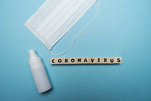 Hygienische Gesichtsmaske mit weißer Flasche Händedesinfektionsmittel mit Coronavirus Wort auf Holzblock isoliert über blauem Hintergrund geschrieben. Konzept zur Prävention von Virusausbrüchen. - Foto, Bild
