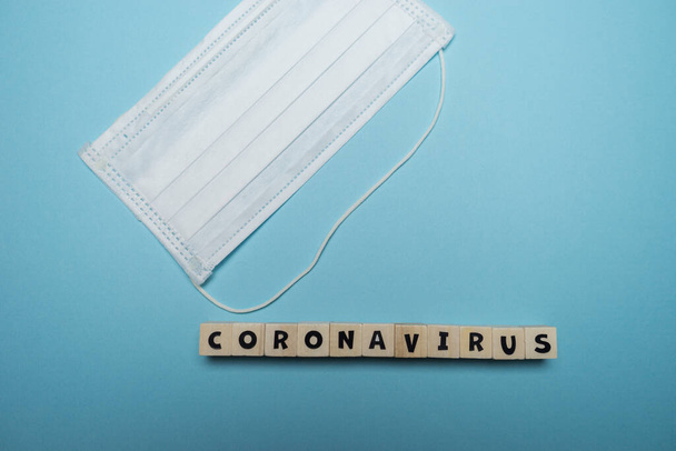 Hygienische Gesichtsmaske mit Coronavirus-Wort auf Holzblock isoliert über blauem Hintergrund geschrieben. Konzept zur Prävention von Virusausbrüchen. - Foto, Bild