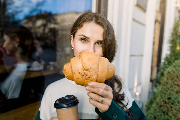 Young pretty stylish woman hiding half of her face with fresh delicious croissant and enjoying lunch break with coffee, standing oudoors near the city building or cafe - Fotografie, Obrázek