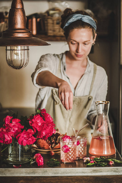 Young woman in linen apron adding ingredients to rose lemonade with ice in glasses over concrete kitchen counter with fresh pink rose flowers on it. Summer refreshing cold beverage drink concept - Foto, afbeelding