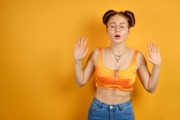 A red-haired girl in an orange top and glasses stands on a yellow background, closing eyes and raising her hands, palms forward - Fotó, kép