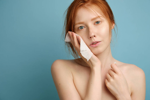Red-haired girl with clean radiant skin and blue eyes, without makeup, presses a napkin to face, standing on a blue background - Fotografie, Obrázek