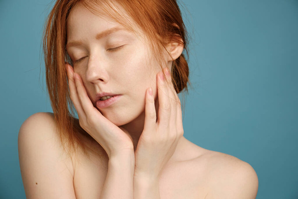 A red-haired girl with clean radiant skin, without makeup, with eyes closed, stands on a blue background, putting palms to face - Fotografie, Obrázek