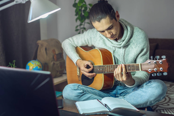 Jovem músico do sexo masculino está aprendendo a tocar guitarra acústica em uma lição on-line usando laptop, à noite pela luz de uma lâmpada em um quarto em casa
.. - Foto, Imagem