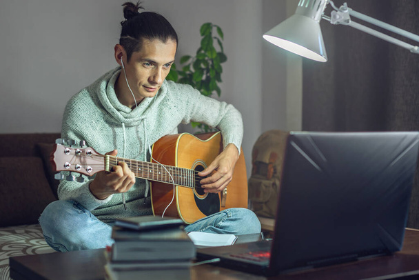 Młody muzyk uczy się grać na gitarze akustycznej w lekcji online, używając laptopa w nocy przy świetle lampy w pokoju w domu.. - Zdjęcie, obraz