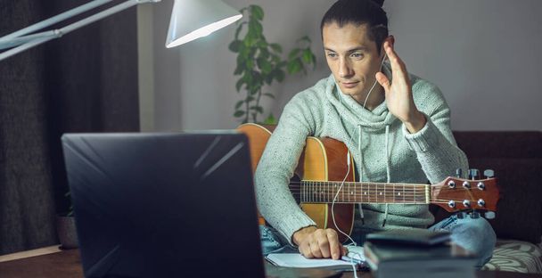 Giovane musicista di sesso maschile sta imparando a suonare la chitarra acustica in una lezione online utilizzando il computer portatile, di notte alla luce di una lampada in una stanza di casa.. - Foto, immagini