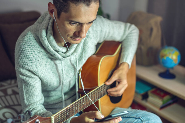 Ένας νεαρός μουσικός μαθαίνει να παίζει ακουστική κιθάρα. Ένας άντρας παίζει κιθάρα από κοντά. - Φωτογραφία, εικόνα