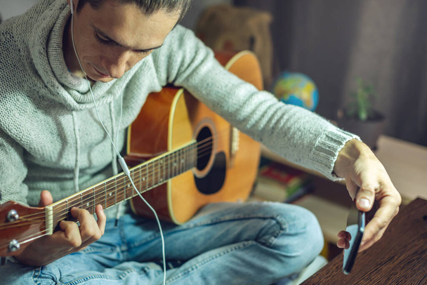 Un jeune musicien apprend à jouer de la guitare acoustique dans une leçon en ligne à l'aide d'une application téléphonique. Un homme étudie des cours à la maison dans une pièce à la lumière d'une lampe - Photo, image