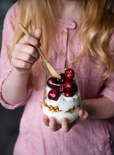 la niña sostiene en su mano una copa con un postre de cerezas rojas frescas y dulces, granola y yogur natural. Sin rostro
 - Foto, Imagen