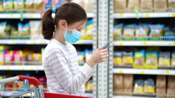 Addicted girl in medical mask playing on smartphone while mother shopping - Záběry, video